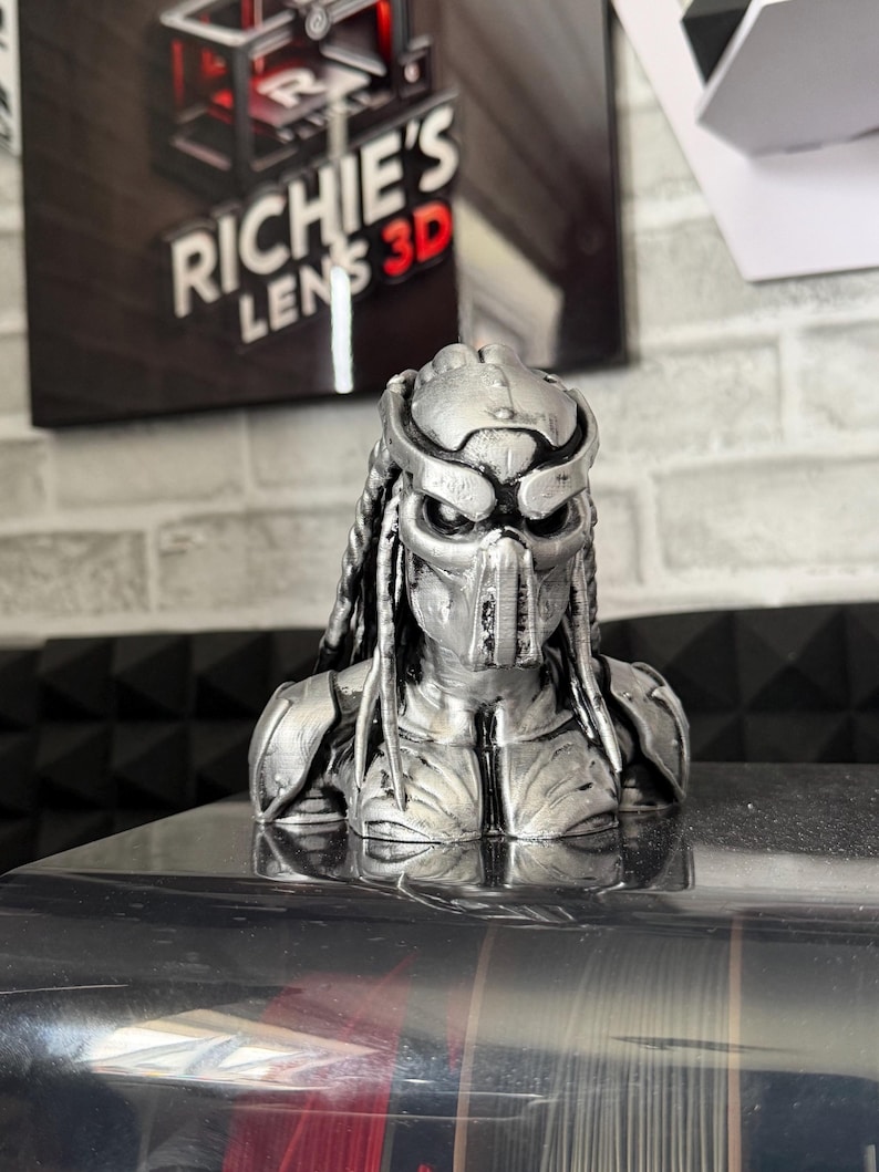 May include: A silver-colored Predator bust, a detailed 3D-printed sculpture. The bust features the character's iconic mask, dreadlocks, and armor. The background includes a sign that reads "RICHIE'S LENS 3D".