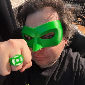 Puede incluir: Una m&aacute;scara y un anillo verdes con un emblema blanco. El anillo est&aacute; en el dedo de una persona. La m&aacute;scara cubre los ojos y la frente. El anillo y la m&aacute;scara son verdes. La persona lleva una camiseta negra con texto blanco.