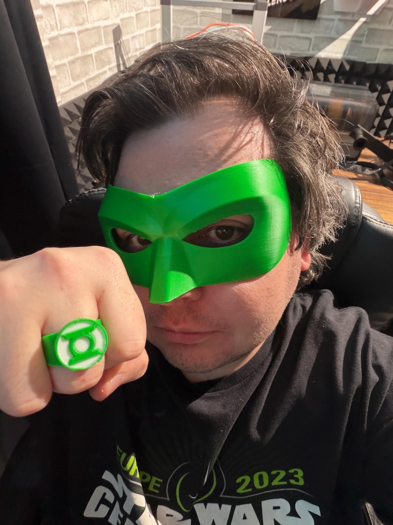 Puede incluir: Una m&aacute;scara y un anillo verdes con un emblema blanco. El anillo est&aacute; en el dedo de una persona. La m&aacute;scara cubre los ojos y la frente. El anillo y la m&aacute;scara son verdes. La persona lleva una camiseta negra con texto blanco.