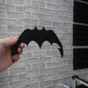 May include: A matte black, bat-shaped object with a symmetrical, pointed wing design. The item features a central vertical structure and sharp, curved edges on both sides, resembling a stylized bat silhouette, held in a neutral position.