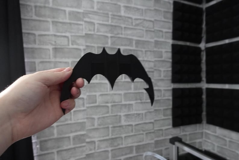 May include: A matte black, bat-shaped object with a symmetrical, pointed wing design. The item features a central vertical structure and sharp, curved edges on both sides, resembling a stylized bat silhouette, held in a neutral position.