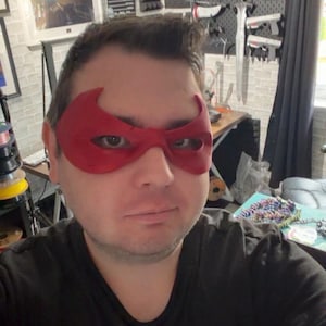 May include: A red superhero mask with a distinct winged shape covering the eye area. The mask features two large, almond-shaped eye openings and a smooth, matte finish, designed to fit across the bridge of the nose and the upper cheeks.