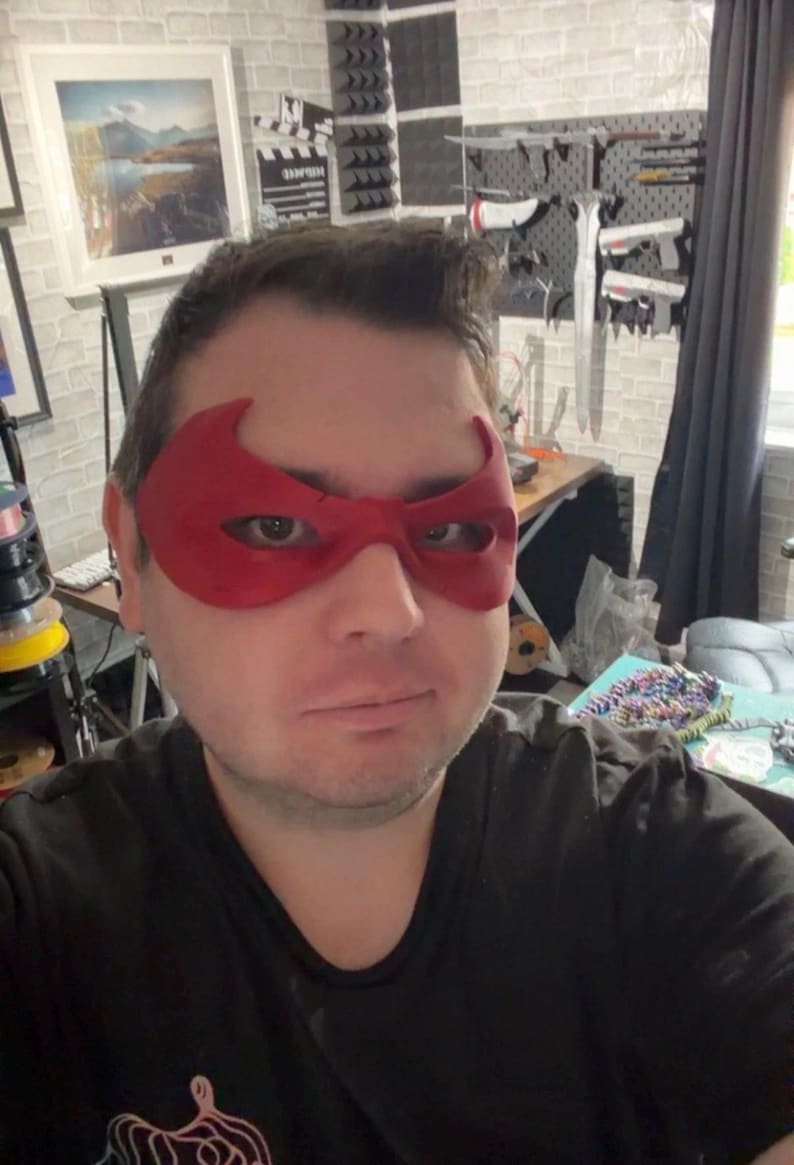 May include: A red superhero mask with a distinct winged shape covering the eye area. The mask features two large, almond-shaped eye openings and a smooth, matte finish, designed to fit across the bridge of the nose and the upper cheeks.
