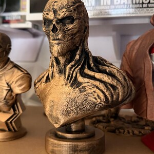May include: A bronze-colored bust of a character with a skeletal face and textured skin. The bust is mounted on a circular base and appears to be a collectible item. The figure has a stern expression and is part of a collection.