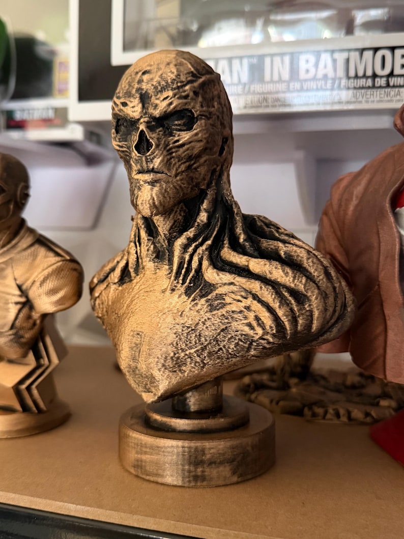May include: A bronze-colored bust of a character with a skeletal face and textured skin. The bust is mounted on a circular base and appears to be a collectible item. The figure has a stern expression and is part of a collection.