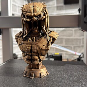 May include: A detailed bronze-colored bust of the Predator alien, featuring intricate armor and dreadlocks. The sculpture showcases the creature's menacing face with open jaws and detailed facial features. The bust is mounted on a decorative base.