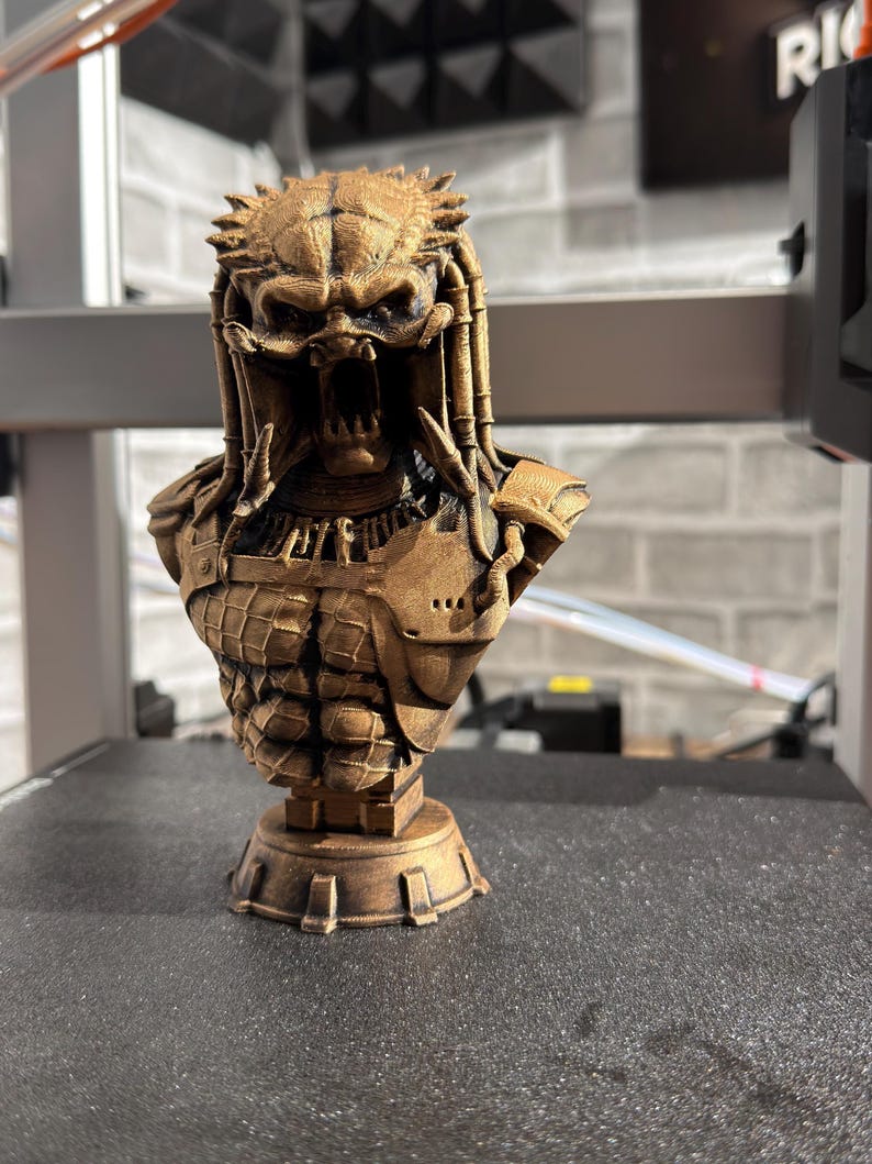 May include: A detailed bronze-colored bust of the Predator alien, featuring intricate armor and dreadlocks. The sculpture showcases the creature's menacing face with open jaws and detailed facial features. The bust is mounted on a decorative base.