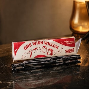 May include: A rectangular red and white box labeled ONE WISH WILLOW with illustrations of two faces and the text AMAZE YOUR FRIENDS and You only get ONE WISH. In front of the box lies a black, elongated, lattice-structured object with hollow openings.