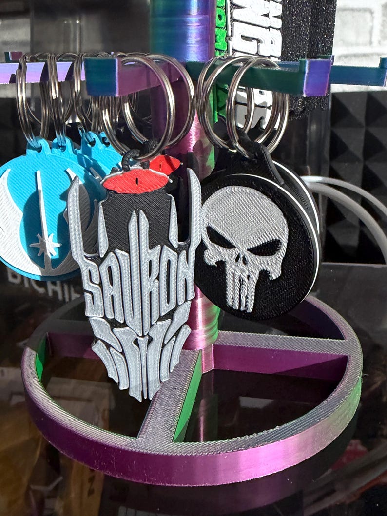K&ouml;nnte beinhalten: Eine Pr&auml;sentation von Schl&uuml;sselanh&auml;ngern mit verschiedenen Designs. Ein Schl&uuml;sselanh&auml;nger zeigt das Wort "SAURON" in einer stilisierten Schriftart, ein anderer hat einen Sch&auml;del und ein dritter ein blaues Design. Die Schl&uuml;sselanh&auml;nger sind an silbernen Ringen befestigt.