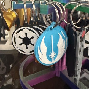 K&ouml;nnte beinhalten: Eine Sammlung von Schl&uuml;sselanh&auml;ngern mit verschiedenen Designs, darunter ein blau-wei&szlig;es Emblem, ein schwarz-wei&szlig;es kreisf&ouml;rmiges Emblem und andere geometrische Formen. Die Schl&uuml;sselanh&auml;nger sind an Metallringen befestigt und auf einem mehrfarbigen St&auml;nder ausgestellt.