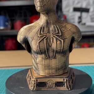 May include: A bronze-colored Spider-Man bust, featuring the character's mask and torso. The bust has a textured, web-like pattern and a large spider emblem on the back. It sits on a black base, showcasing the superhero's iconic design.