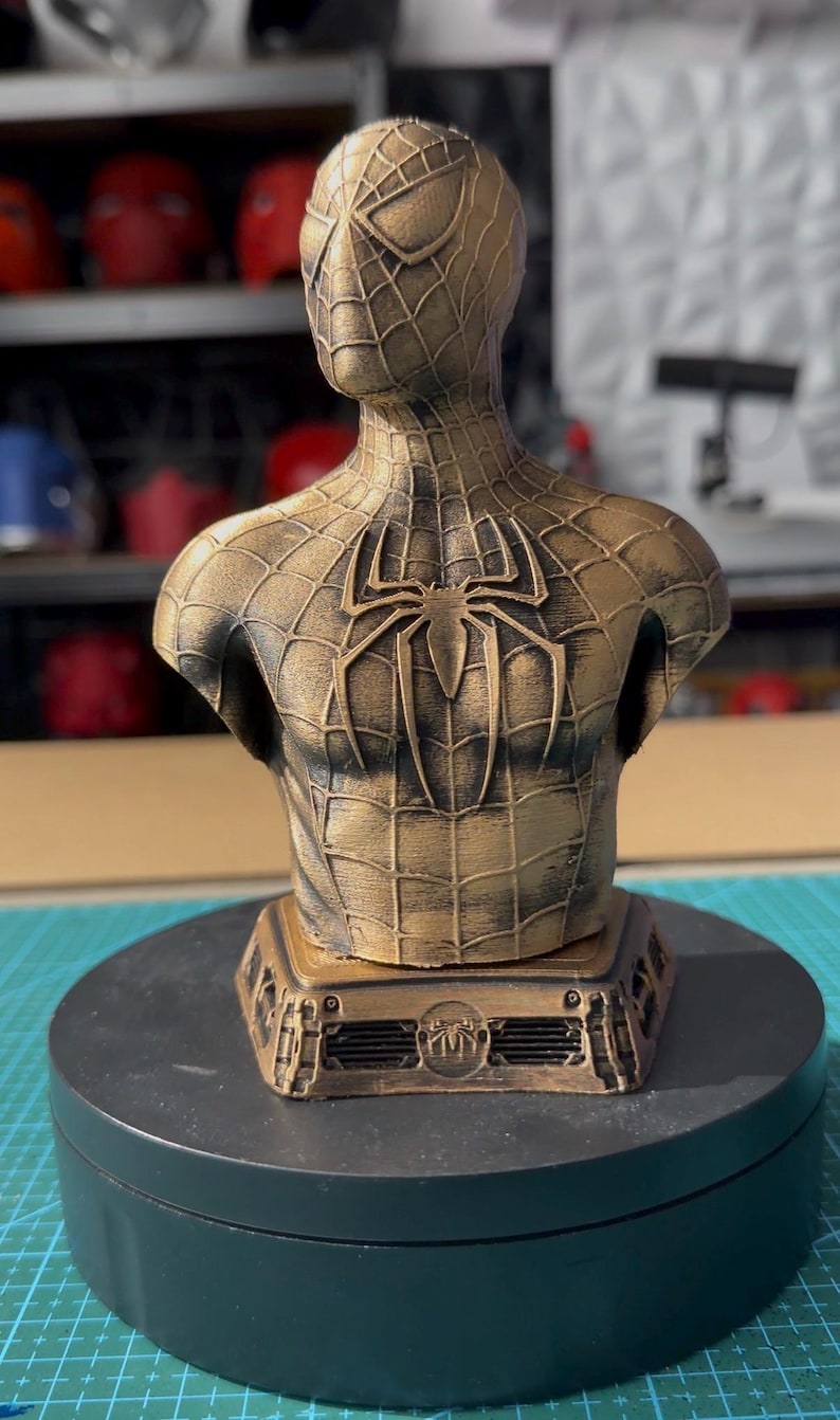 May include: A bronze-colored Spider-Man bust, featuring the character's mask and torso. The bust has a textured, web-like pattern and a large spider emblem on the back. It sits on a black base, showcasing the superhero's iconic design.