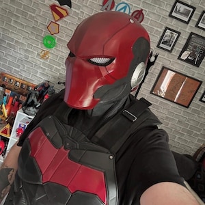 May include: A Red Hood helmet and chest armour set. The helmet is mainly red with a black trim and mesh eye coverings. The chest armour is black with a red bat symbol. The design is reminiscent of a comic book superhero.