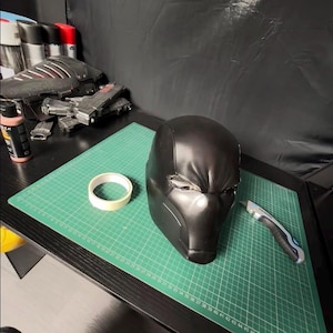 Op de afbeelding: Een zwarte helm in futuristische stijl met een matte afwerking. De helm ligt op een groene snijmat, naast een rol tape en een hobbymes. Andere knutselspullen en een model zijn zichtbaar op de achtergrond.