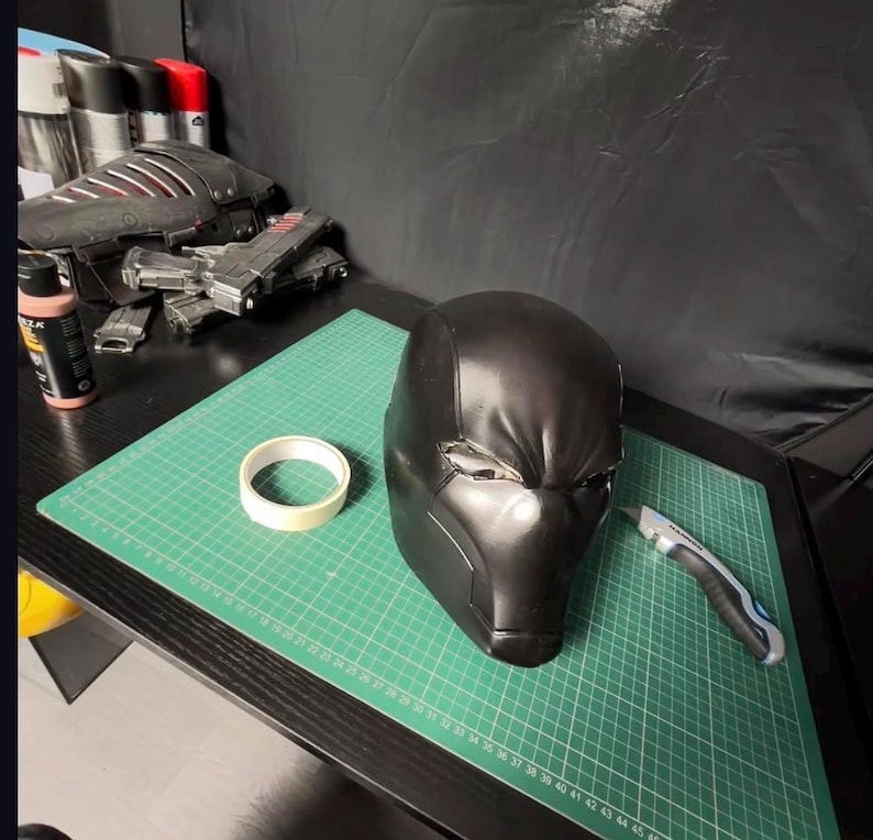 Op de afbeelding: Een zwarte helm in futuristische stijl met een matte afwerking. De helm ligt op een groene snijmat, naast een rol tape en een hobbymes. Andere knutselspullen en een model zijn zichtbaar op de achtergrond.