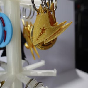 K&ouml;nnte beinhalten: Eine Pr&auml;sentation goldfarbener Schl&uuml;sselanh&auml;nger, die jeweils an einem silbernen Schl&uuml;sselring befestigt sind. Die Schl&uuml;sselanh&auml;nger weisen ein Sterndesign auf und werden auf einem wei&szlig;en, mehrstufigen St&auml;nder ausgestellt. Weitere blaue und goldene Schl&uuml;sselanh&auml;nger sind ebenfalls sichtbar.