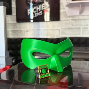 Puede incluir: Una m&aacute;scara de superh&eacute;roe verde brillante y un anillo a juego con detalles dorados. El anillo presenta un emblema verde. La m&aacute;scara y el anillo parecen estar impresos en 3D. El fondo incluye un letrero que dice "Richie's Lens 3D".