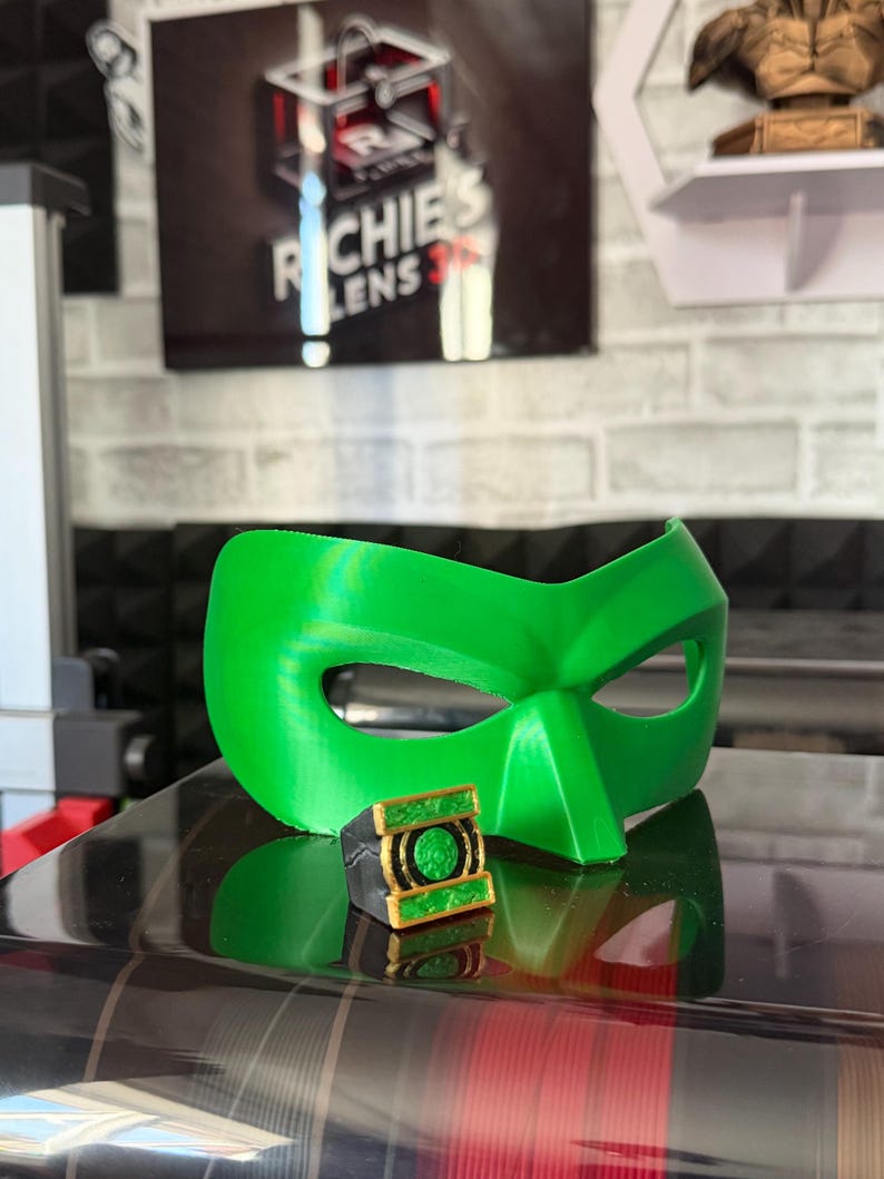 Puede incluir: Una m&aacute;scara de superh&eacute;roe verde brillante y un anillo a juego con detalles dorados. El anillo presenta un emblema verde. La m&aacute;scara y el anillo parecen estar impresos en 3D. El fondo incluye un letrero que dice "Richie's Lens 3D".