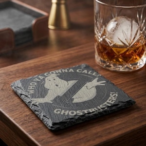 May include: A square, dark gray slate coaster with the Ghostbusters logo and the text "WHO YA GONNA CALL? GHOSTBUSTERS!". A glass of amber liquid with a large ice sphere sits nearby on a wooden surface.
