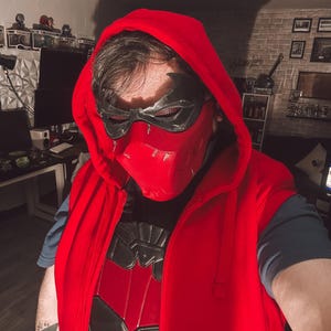 May include: A red hooded vest with a black and red chest plate. The person is wearing a black and red mask with a hood. The outfit is a cosplay costume.