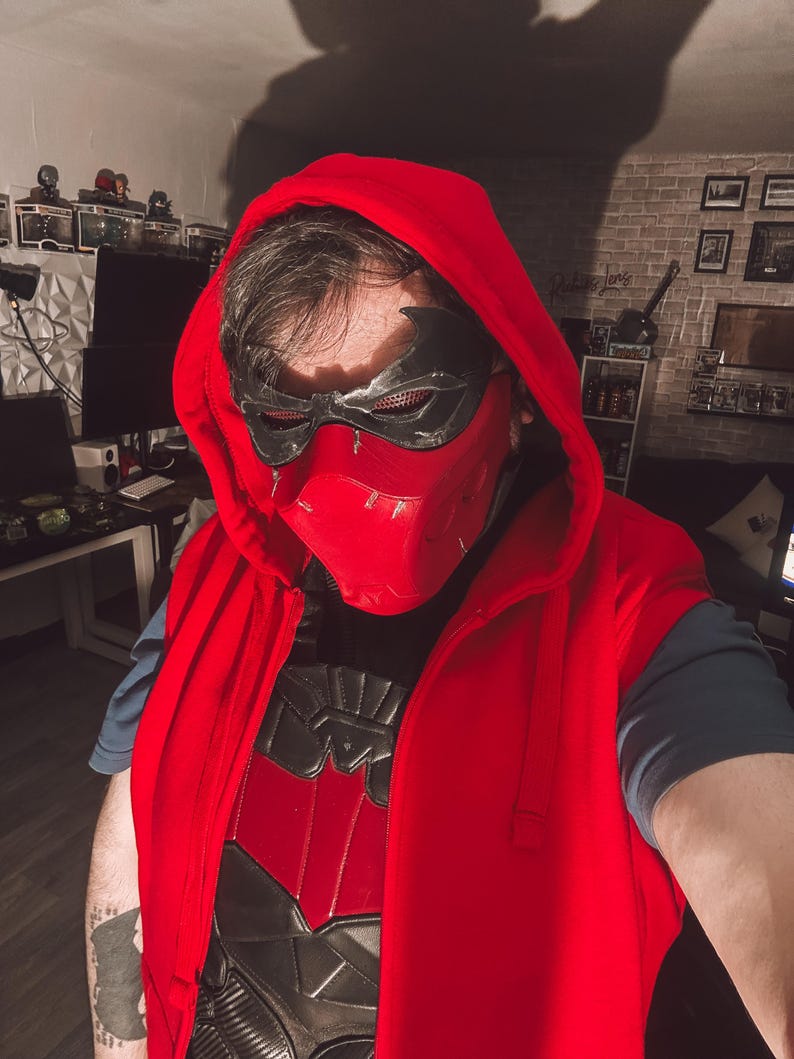 May include: A red hooded vest with a black and red chest plate. The person is wearing a black and red mask with a hood. The outfit is a cosplay costume.