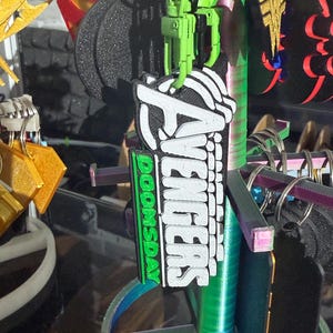 K&ouml;nnte beinhalten: Eine Pr&auml;sentation von Schl&uuml;sselanh&auml;ngern mit verschiedenen Designs. Der Hauptschl&uuml;sselanh&auml;nger zeigt das Wort "AVENGERS" in Wei&szlig;, mit "DOOMSDAY" in Gr&uuml;n. Weitere Schl&uuml;sselanh&auml;nger sind ein goldener Stern, ein schwarzer Reifen und ein rot-gelbes Emblem. Die Schl&uuml;sselanh&auml;nger sind an silbernen Schl&uuml;sselringen befestigt.