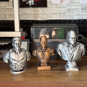 May include: Three superhero bust statues. The Punisher bust is silver with a detailed chest plate. The Daredevil bust is bronze with a "D" emblem. The third bust is silver and depicts a man in a suit jacket. All are on a wooden surface.