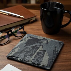 May include: A black and gray slate coaster featuring a Batman comic book design. The coaster is square and sits on a wooden surface, next to a black mug, glasses, and a notebook. The text "Gotham Nights" is visible.