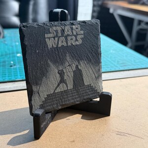 May include: A black slate coaster with the words "STAR WARS" etched into the surface. The coaster features a silhouette of a lightsaber duel. The coaster is displayed on a black stand.