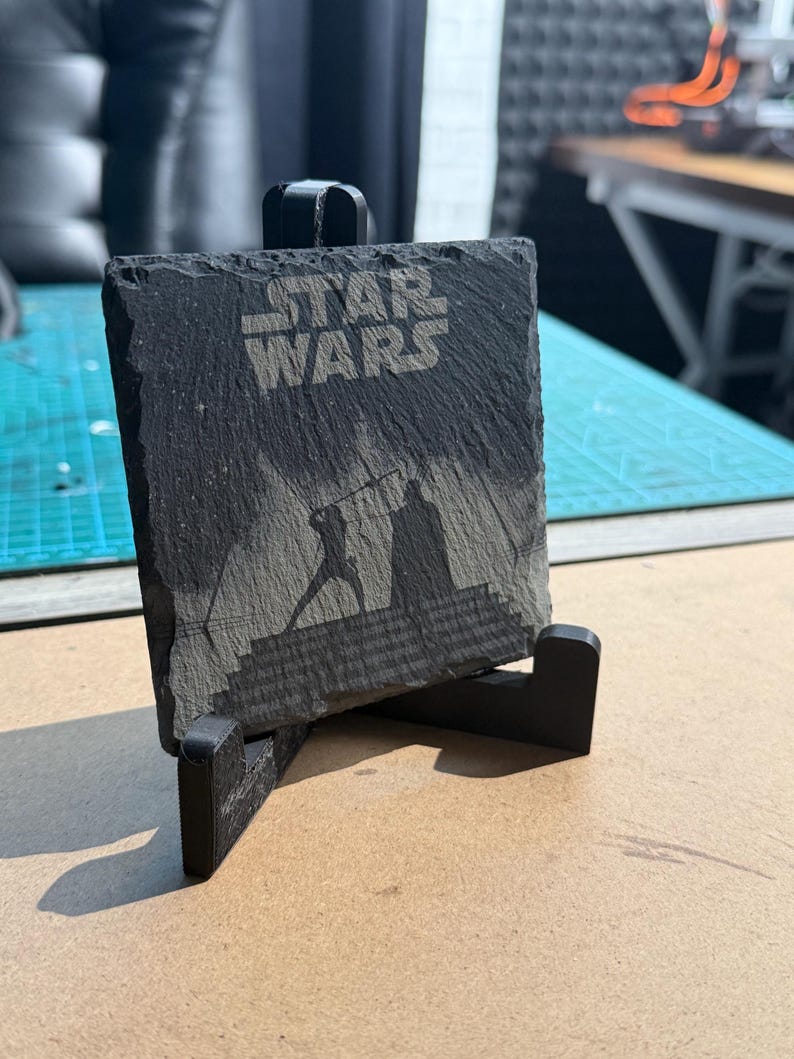 May include: A black slate coaster with the words "STAR WARS" etched into the surface. The coaster features a silhouette of a lightsaber duel. The coaster is displayed on a black stand.