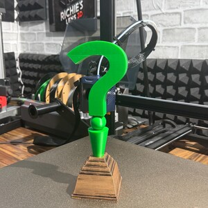 May include: A 3D-printed green question mark on a brown, tiered base. The question mark is the main feature, with a smooth, bright green finish. The base has a wood-like texture. The item is displayed on a flat surface, with a 3D printer in the background.
