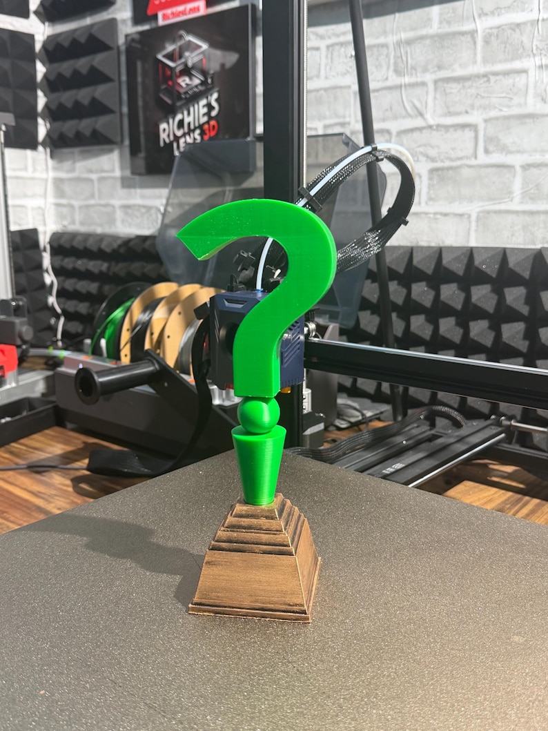 May include: A 3D-printed green question mark on a brown, tiered base. The question mark is the main feature, with a smooth, bright green finish. The base has a wood-like texture. The item is displayed on a flat surface, with a 3D printer in the background.
