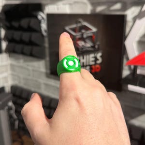 Puede incluir: Un anillo verde con un emblema circular blanco, usado en un dedo. El anillo presenta un dise&ntilde;o detallado y probablemente sea un art&iacute;culo hecho por un fan. El fondo incluye un letrero con el texto "Sophie's 3D".