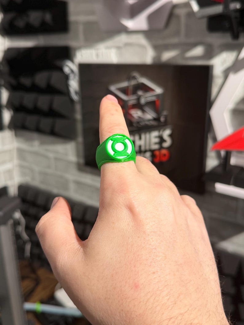 Puede incluir: Un anillo verde con un emblema circular blanco, usado en un dedo. El anillo presenta un dise&ntilde;o detallado y probablemente sea un art&iacute;culo hecho por un fan. El fondo incluye un letrero con el texto "Sophie's 3D".