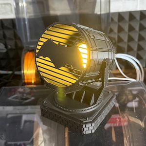 以下が含まれることがあります： 黒い3Dプリントのスポットライト風ランプ。コウモリのシルエットが特徴です。ランプは内側から黄色の光を放ち、正方形の台座に取り付けられています。サーチライトを模したデザインです。