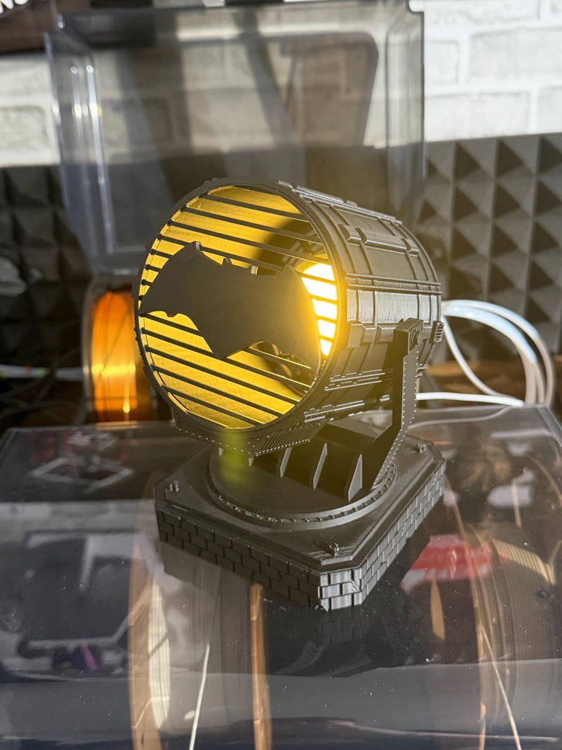 以下が含まれることがあります： 黒い3Dプリントのスポットライト風ランプ。コウモリのシルエットが特徴です。ランプは内側から黄色の光を放ち、正方形の台座に取り付けられています。サーチライトを模したデザインです。