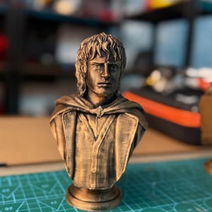 May include: A bronze-colored bust of a man with shoulder-length hair and a button-down shirt. The bust has a detailed face and is mounted on a round base. The figure is wearing a cloak. The bust is likely a collectible or decorative item.