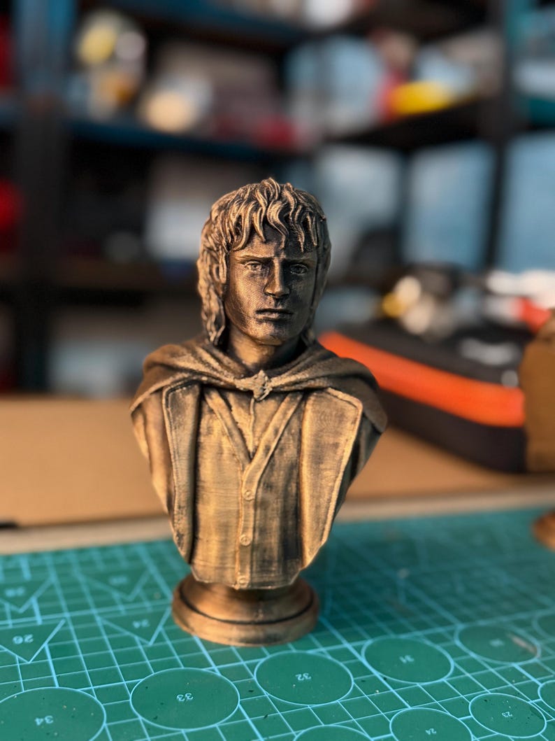 May include: A bronze-colored bust of a man with shoulder-length hair and a button-down shirt. The bust has a detailed face and is mounted on a round base. The figure is wearing a cloak. The bust is likely a collectible or decorative item.