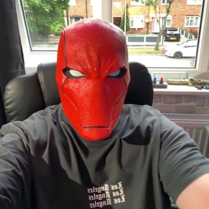 Op de afbeelding: Een felrood masker met witte oogopeningen. Het masker bedekt het hoofd en heeft een textuur. De persoon draagt een donkergrijs t-shirt met de tekst "Los Angeles" erop.