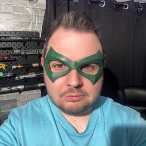 May include: A green superhero-style eye mask with angular, pointed edges covering the eye area. The mask features a smooth, solid green finish and is worn over the face. The wearer is dressed in a light blue t-shirt with a black logo patch.