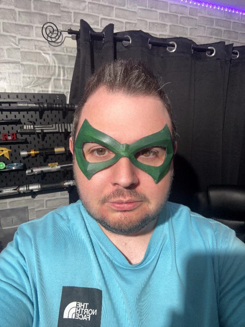 May include: A green superhero-style eye mask with angular, pointed edges covering the eye area. The mask features a smooth, solid green finish and is worn over the face. The wearer is dressed in a light blue t-shirt with a black logo patch.