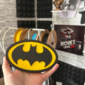 May include: An oval-shaped Batman logo plaque featuring a black bat silhouette centered on a bright yellow background, enclosed by a thick black border. The design is flat and constructed with distinct layers, showcasing a classic comic book emblem.