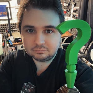 May include: A green question mark trophy with a wooden base. The trophy is held in front of a person wearing a black shirt. The background includes framed artwork and soundproofing panels.
