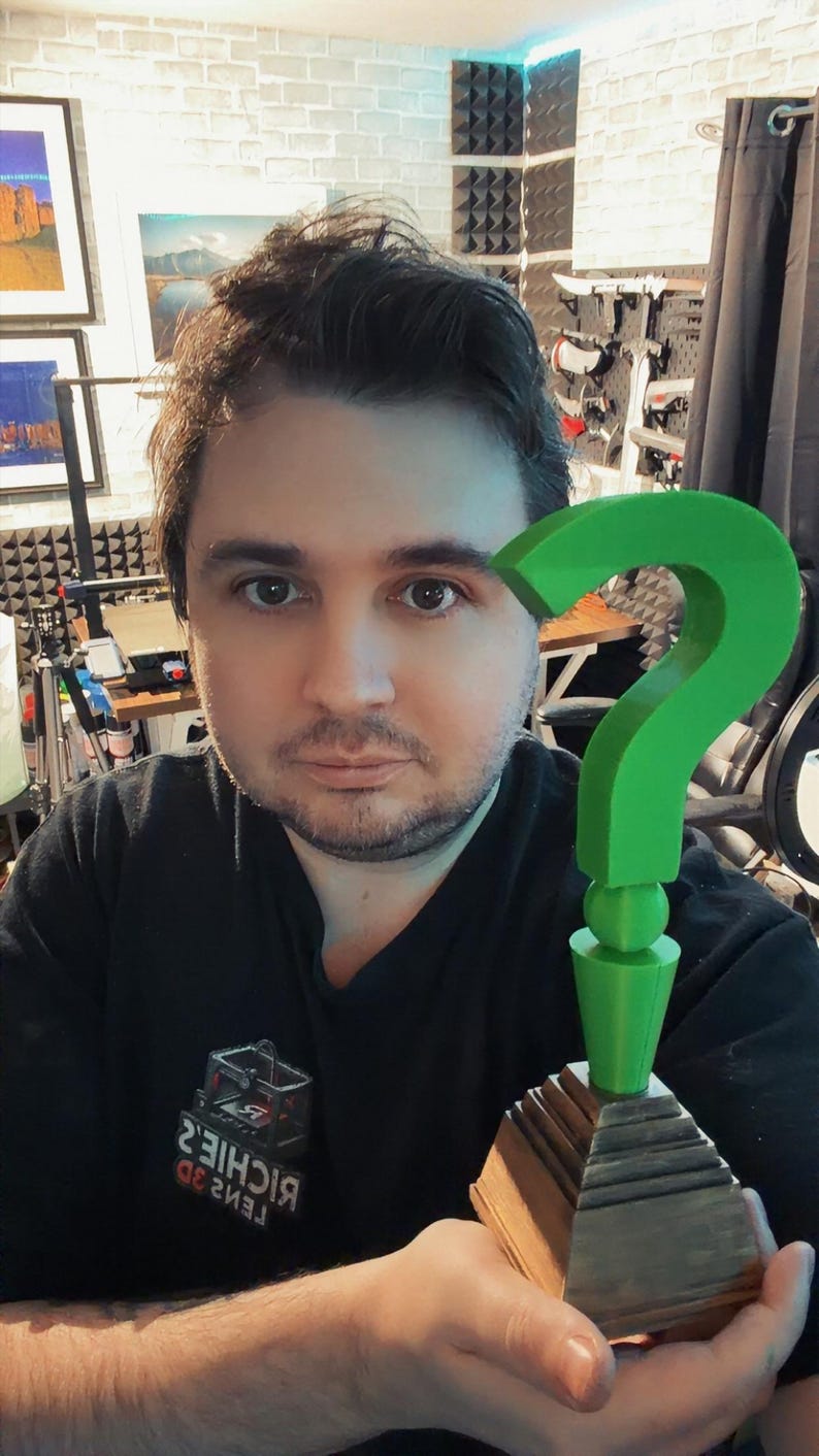 May include: A green question mark trophy with a wooden base. The trophy is held in front of a person wearing a black shirt. The background includes framed artwork and soundproofing panels.