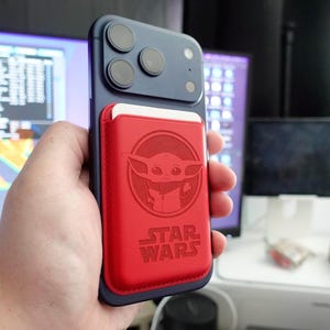 May include: A navy blue smartphone with a red Star Wars phone wallet featuring an engraved image of Grogu. The wallet is attached to the back of the phone. The phone has three camera lenses.