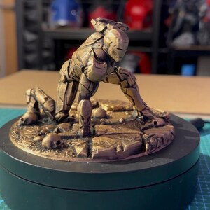 May include: A bronze-colored Iron Man figurine in a kneeling pose, set on a circular base. The figure is highly detailed, with visible armor plating and a helmet. The base features a textured surface with small skull details.