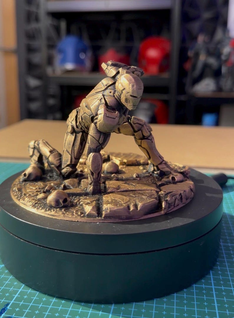May include: A bronze-colored Iron Man figurine in a kneeling pose, set on a circular base. The figure is highly detailed, with visible armor plating and a helmet. The base features a textured surface with small skull details.