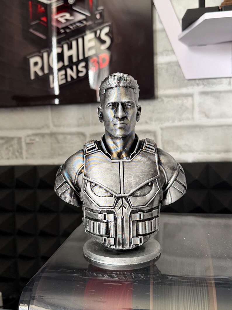 May include: A silver-colored bust of a man wearing a tactical vest with a skull emblem. The bust is 3D-printed and sits on a circular base. The background includes a sign that reads "Richie's Lens 3D".