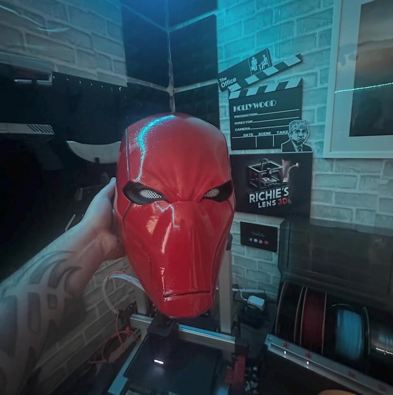 Op de afbeelding: Een glanzende rode Red Hood-helm met witte mesh oogopeningen. Het masker wordt vastgehouden door een hand met een tatoeage. De achtergrond bevat een filmklapper en een bord met "Richie's Lens 3D".