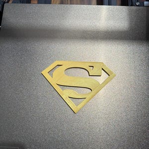 May include: A gold-colored 3D printed Superman logo emblem centered on a dark gray textured build plate. The build plate features a white warning label with the text CAUTION HIGH TEMPERATURE and the brand name ANYCU visible in the corner.